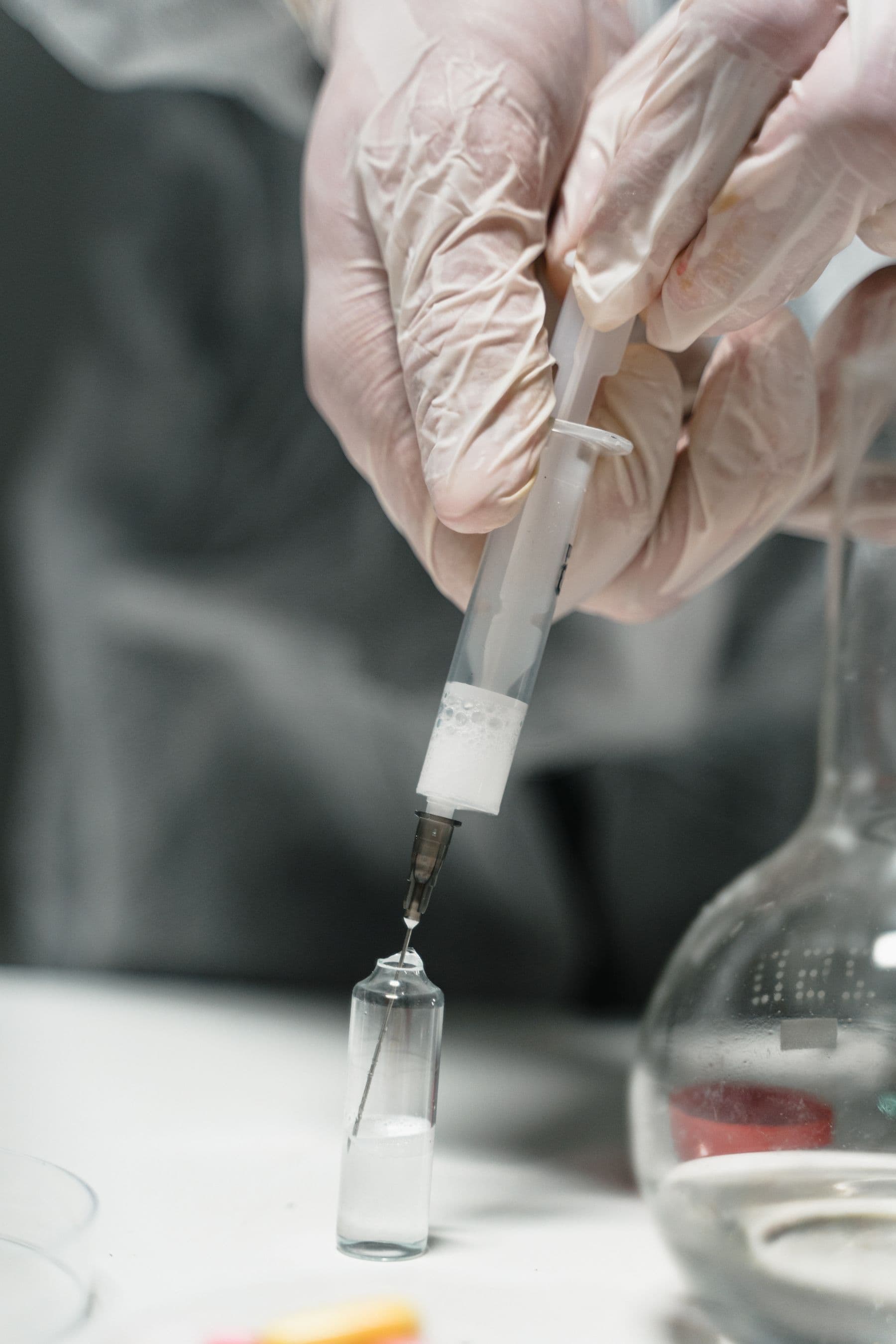 Gloved hand holding a syringe above a small glass vial on a laboratory bench