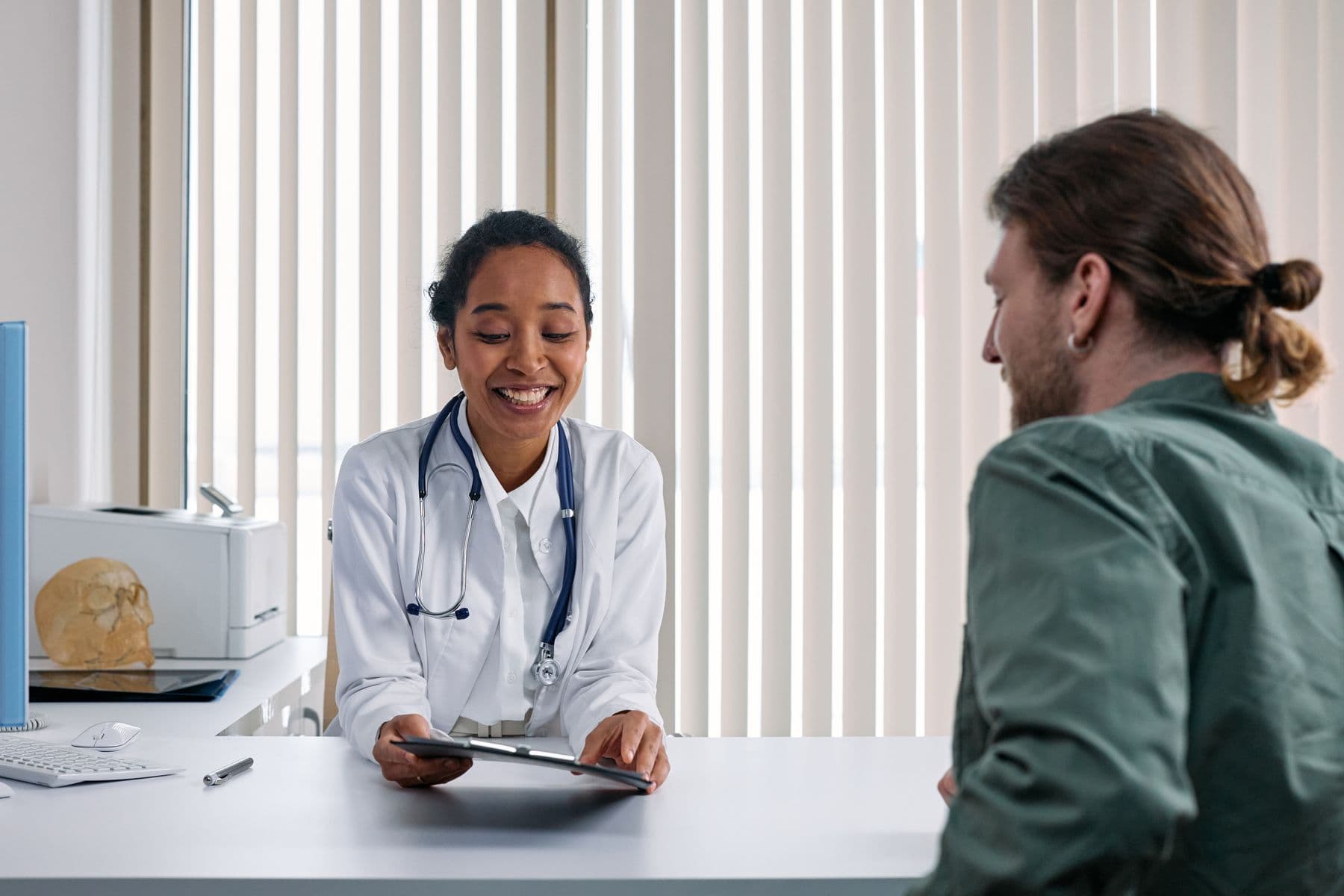 Clinician in a white coat consulting a patient across a desk, clipboard in hand